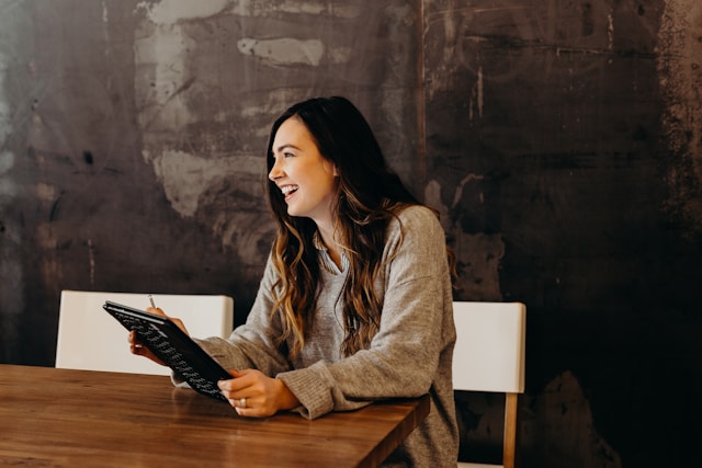 A woman sits at a wooden table, laughing with a tablet in her hand.
