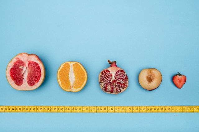 Several differently sized, sliced fruits appear in a line with a measuring tape underneath them against a blue background.