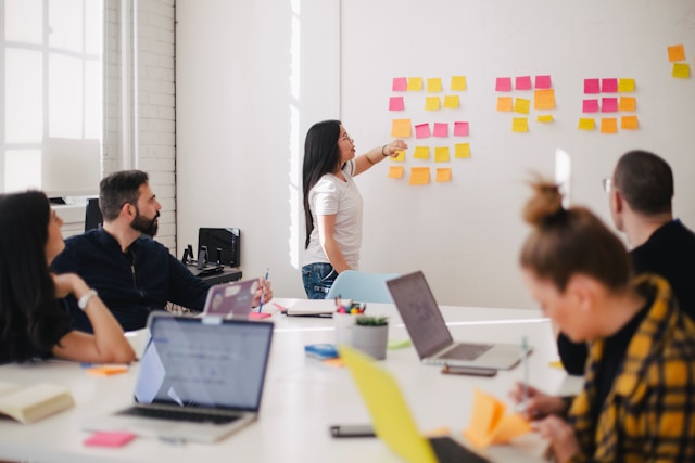 A group of people sit around a table with laptops, looking at a woman standing by a whiteboard and placing sticky notes.