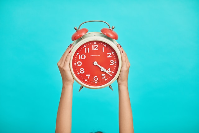 Two hands hold up a red analog alarm clock against a blue background.

