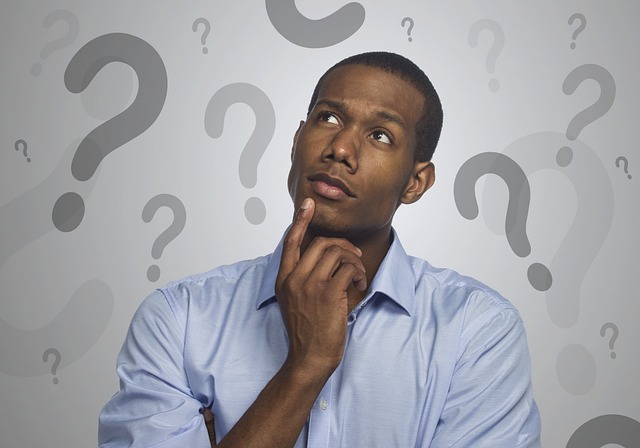 A man stands with a ponderous look on his face against a grey background covered in question marks. 