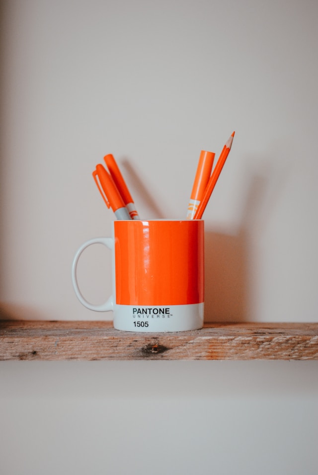 Orange markers in an orange Pantone mug on a wooden shelf.
