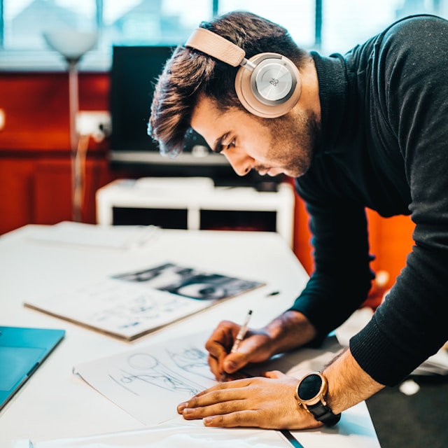 A man wearing headphones leans over a desk, drawing on a piece of paper.
