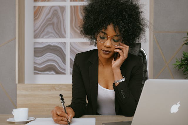 A woman in a black blazer holds a phone to her ear while writing in a notebook. There is a laptop on her desk.
