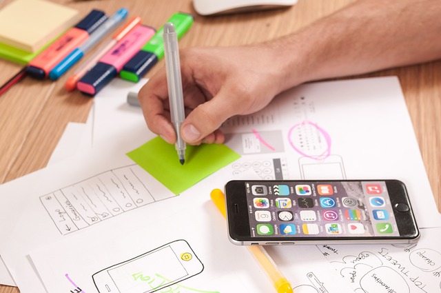 A designer draws a wireframe for a mobile app surrounded by paper and colorful pens. 
