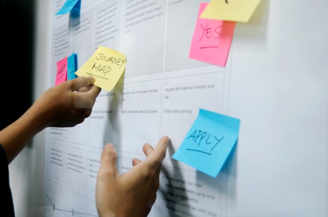 A person holds a yellow sticky note reading "Journey Map" and pins it to a wall.
