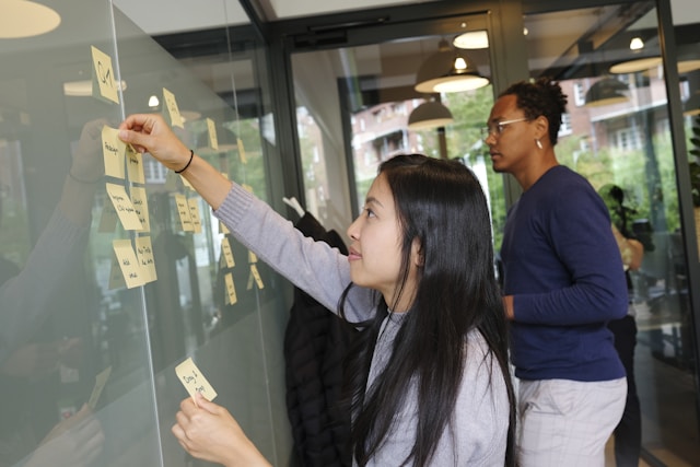 Two people planning a workflow by adding notes to a meeting room’s wall.