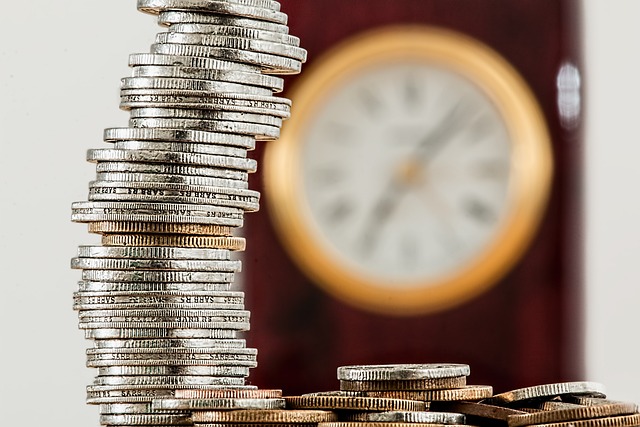 A close-up of a tower of coins in front of an out-of-focus clock dial.
