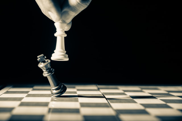 A close-up of a person knocking over a black King chess piece with a white King chess piece. 
