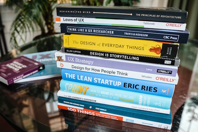 A stack of UX books on a glass-top wooden table.