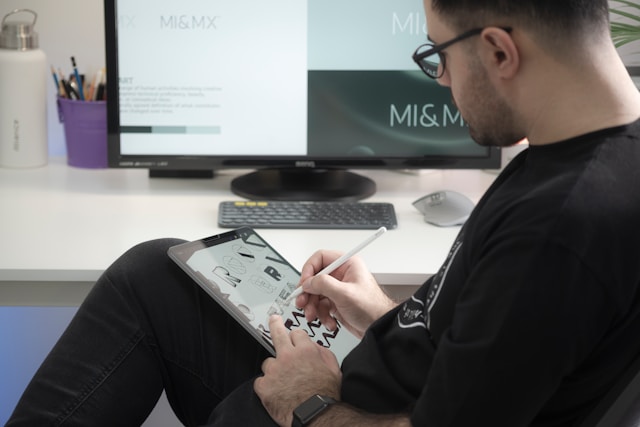 A graphic designer makes illustrations on his smart tablet while sitting in front of a computer.