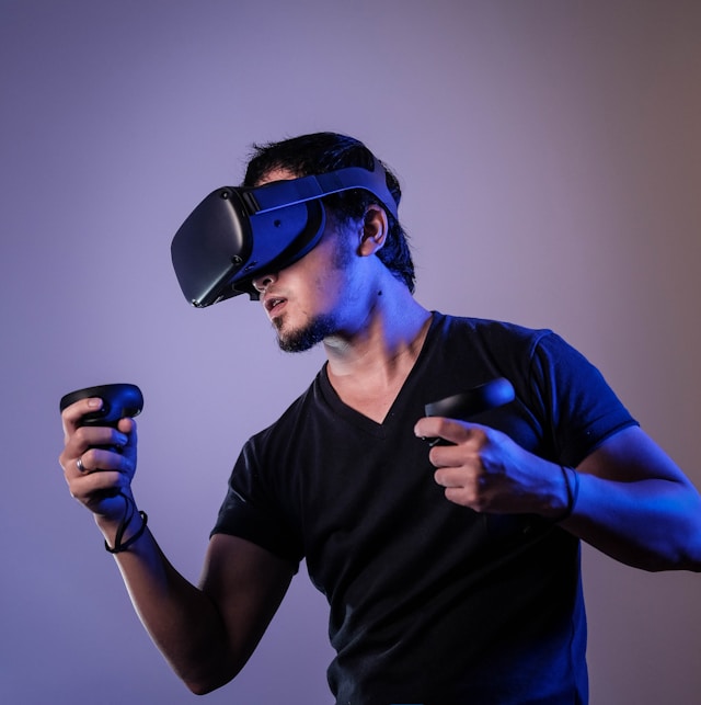A man uses a black VR headset, holding two accompanying devices in each hand.
