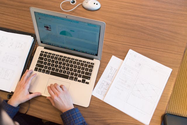 A person works on their laptop, looking at user research data next to a stack of paper wireframes.