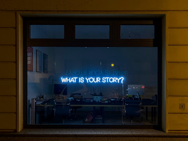 An office window that displays a blue neon sign reading the phrase “What is your story?”