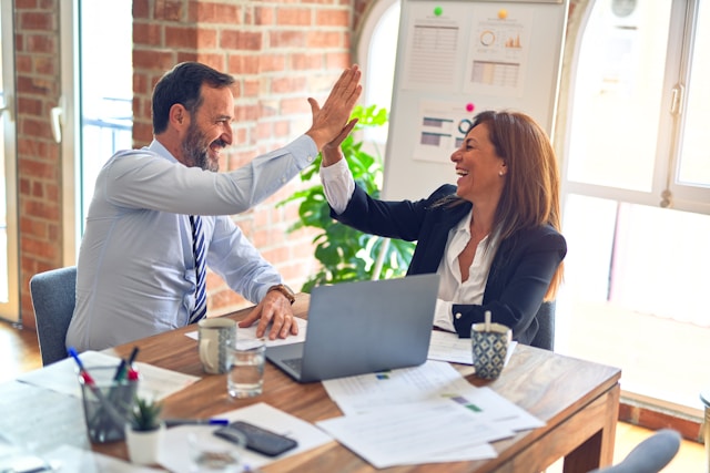 Two co-workers smiling and high-fiving each other as they collaborate over a laptop in an office. 