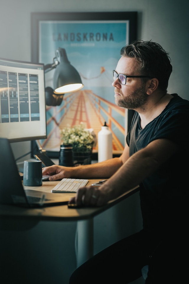 A man in a black crew neck t-shirt and black-framed glasses working in front of a computer.
