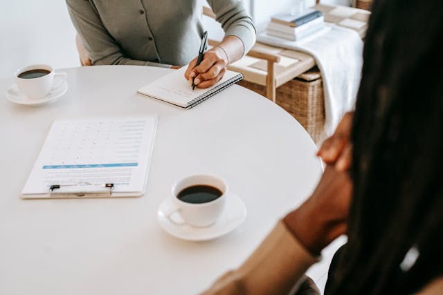 Two people sitting at a table with coffee and a clipboard, while one of them is writing in a notebook.