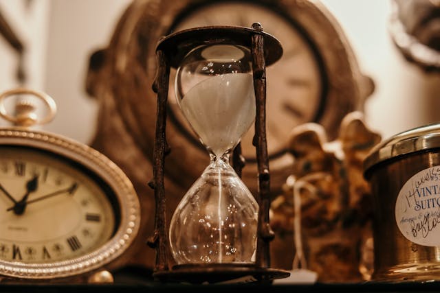 A close-up of a clear hourglass filled with sand and surrounded by clocks 