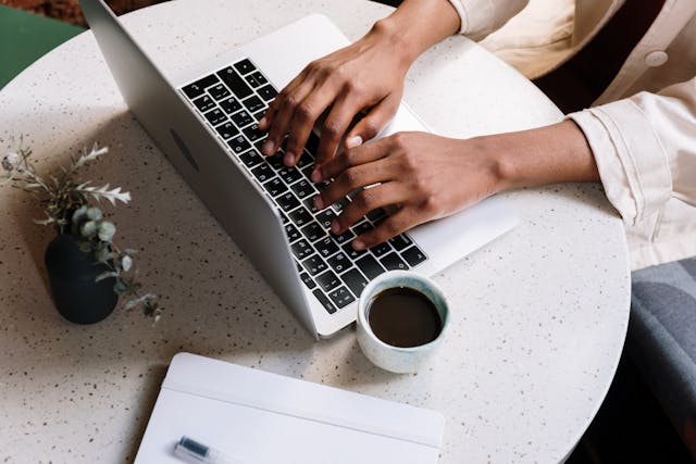 A person typing out content on a laptop with a cup of coffee, a notebook, and a plant next to it.