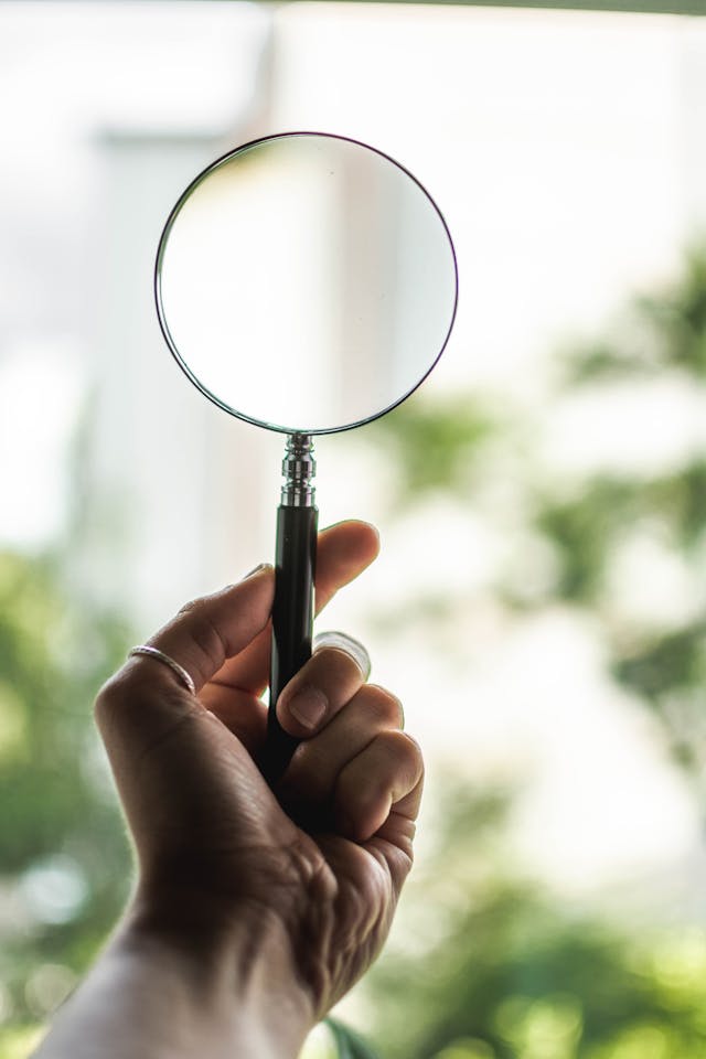 A close-up of a person holding a magnifying glass against an obscure, white and green background. 