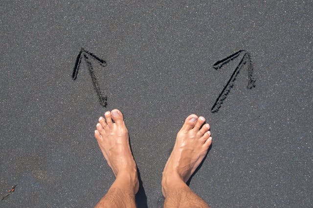 A person stands barefoot on black sand with two arrows in front of them, pointing toward different directions. 