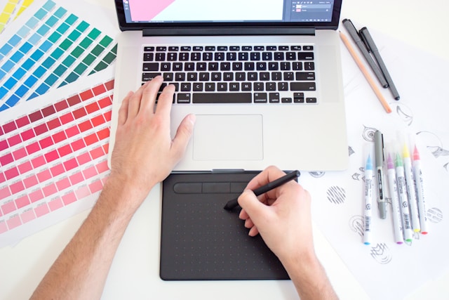 A person using a drawing tablet with a Mac that is surrounded by various color charts.