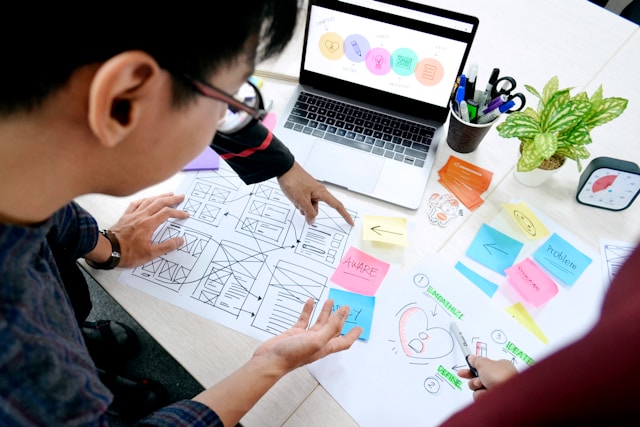 Three people look at papers on a desk, with wireframes and sticky notes dotted around.
