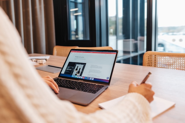 A person sitting at a table using a Macbook while writing in a notebook.