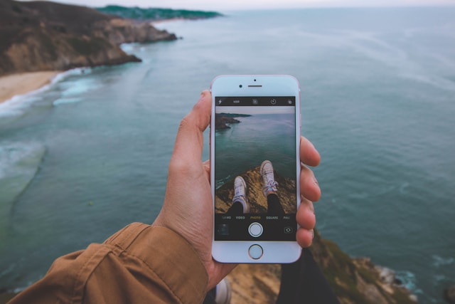 A close-up of a person sitting on top of a hill and holding their phone with the Camera app open. 