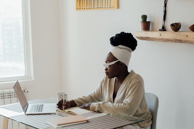 A woman sitting in front of a laptop while at home, making notes in a small notebook. 
