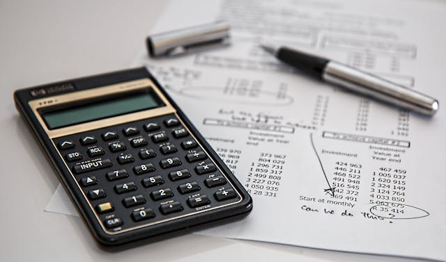 A close-up of a calculator and a pen on top of a piece of paper containing various monetary values. 
