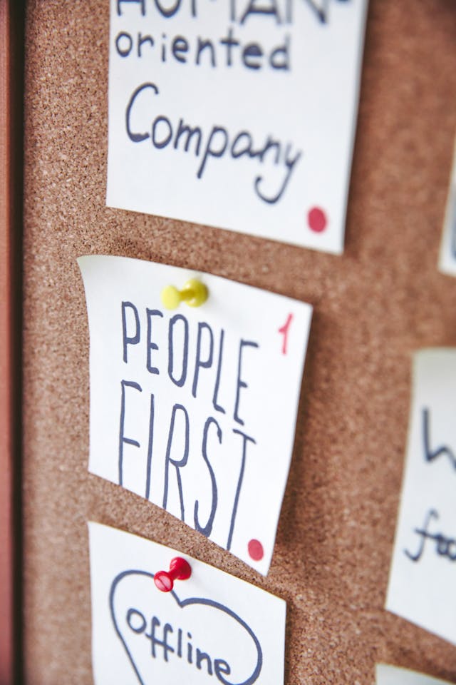 A close-up of small white notes pinned against a brown board. One note reads the phrase ‘People First.’ 
