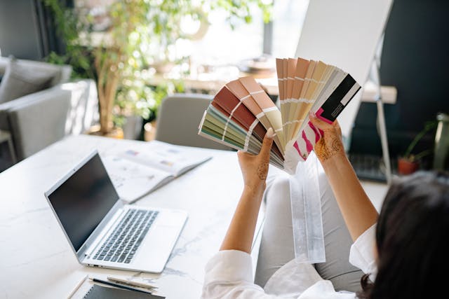 A UI designer holding a color swatch fan beside a white table and silver laptop. 
