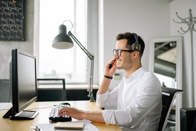 A smiling customer service agent sits at their desk while wearing a headset and looking at their computer. 