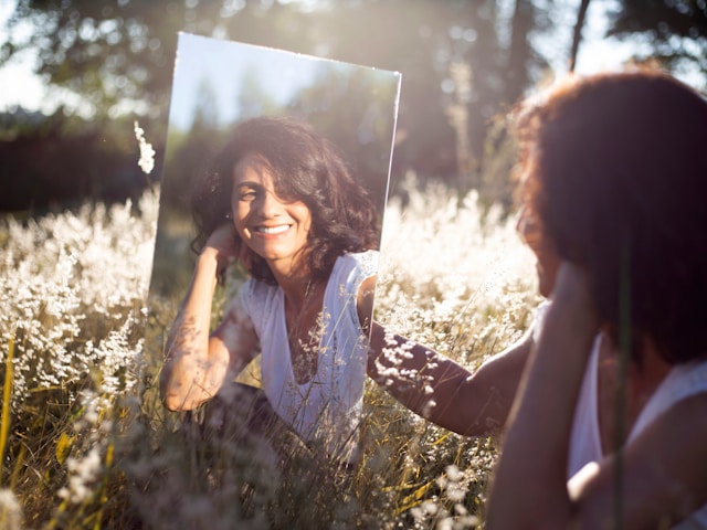 A woman smiling at herself in a mirror while sitting in a sunlit field. 
