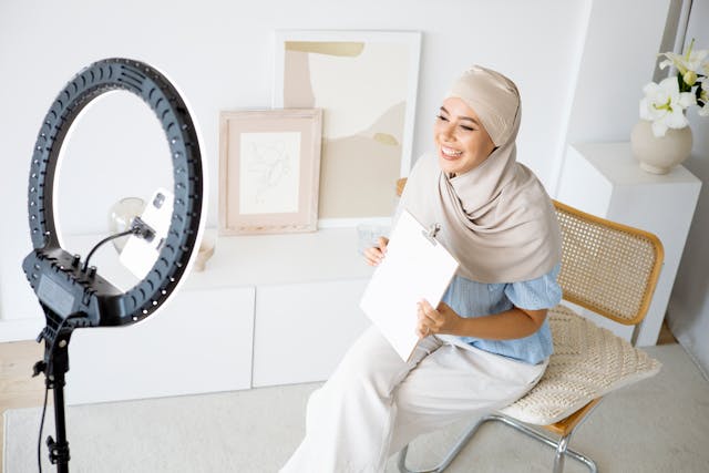 A woman smiles while holding a clipboard and recording a video with her phone and ring light. 
