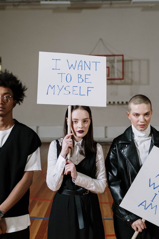 Three students stand in a gym holding signs with the one in the middle reading the phrase “I want to be myself.” 
