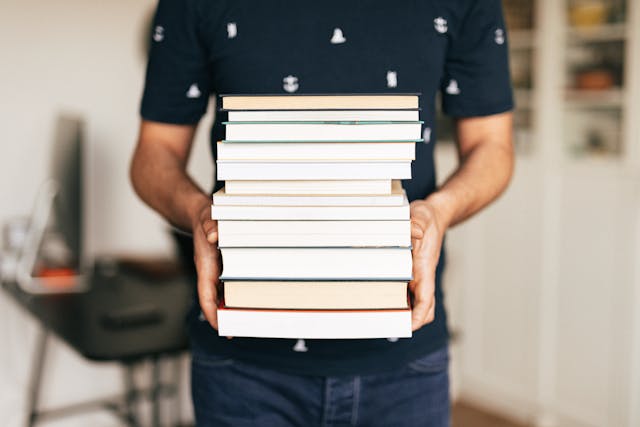 A person wearing a black shirt carrying a tall stack of books. 