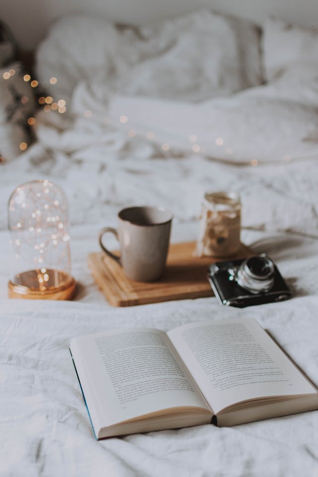 An open book sitting on a white bed near a camera, mug, and wooden tray. 

