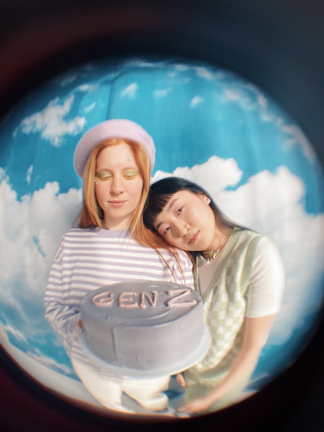A photo taken with a fisheye lens of two brightly dressed teenagers holding a lilac cake that reads the phrase “Gen Z.” 
