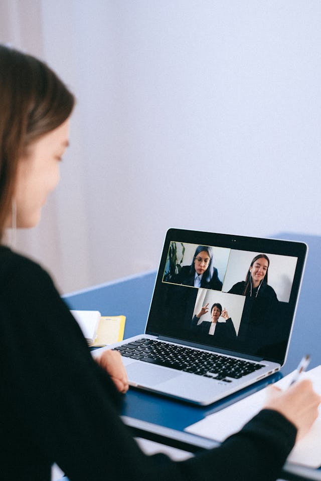 A woman writes notes while participating in a video conference call with three other people.  
