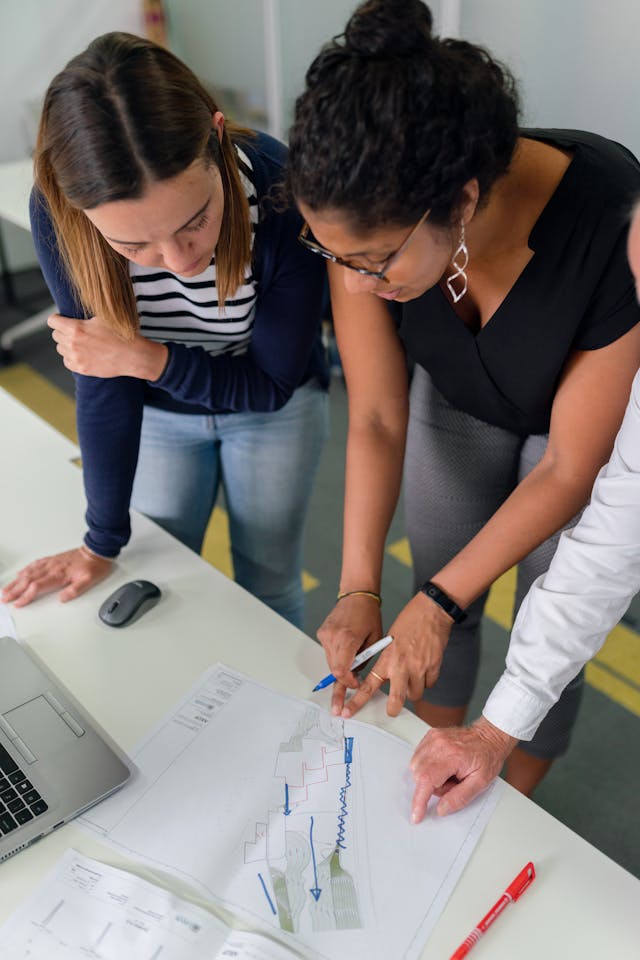 Two co-workers stand over a table looking at a piece of paper, resembling onboarding buddies. 
