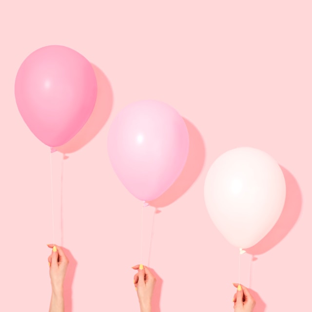 Three people hold up three balloons that display a small range of shades and tints of pink.