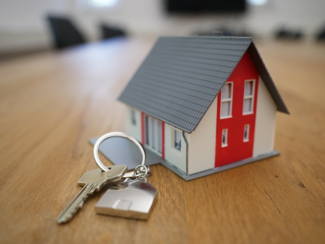  A miniature red and white house sits on a wooden table next to a pair of keys. 

