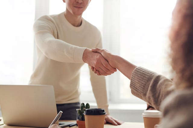 Two people shake hands over a white table during an interview. 
