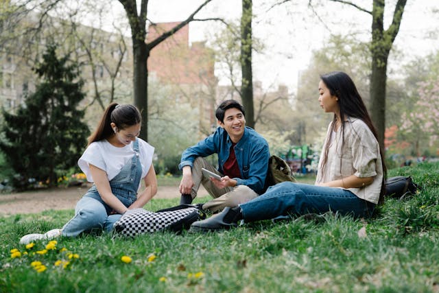 Three students smile and talk to each other while sitting on the grass. 
