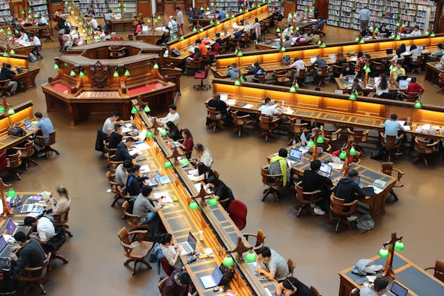 A university library full of students reading books and using their laptops. 
