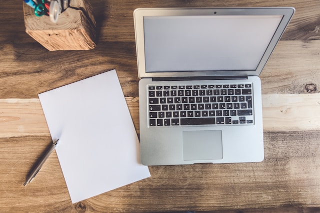 A bird’s-eye view of a blank piece of paper and a pen next to a silver laptop. 
