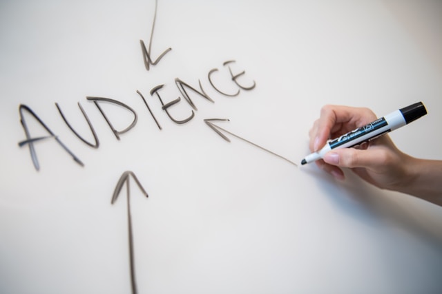 A person draws an arrow on a whiteboard, pointing to the capitalized word “Audience.”
