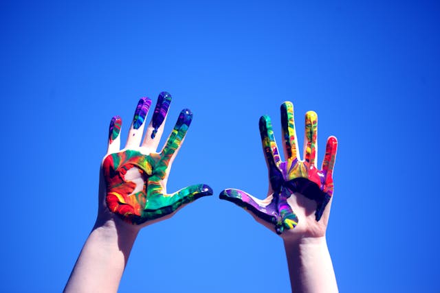 A person holds their colorful, paint-covered hands up to the bright blue sky.